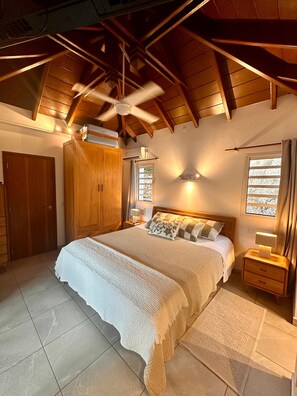 Quarto com cama grande, vigas de madeira no teto, guarda-roupa, mesa de cabeceira com abajur e janela com cortinas.