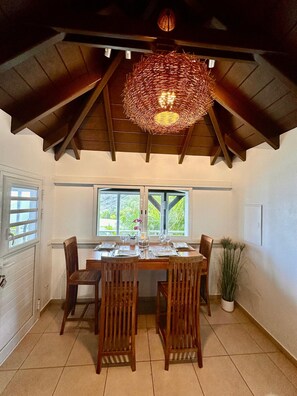 A dining area with a wooden table set for four, surrounded by chairs, under a sloped ceiling with exposed beams. - Villa des Rocher – Private Jacuzzi, Ocean View, and Serenity in Saint Barth (Grand Fond)