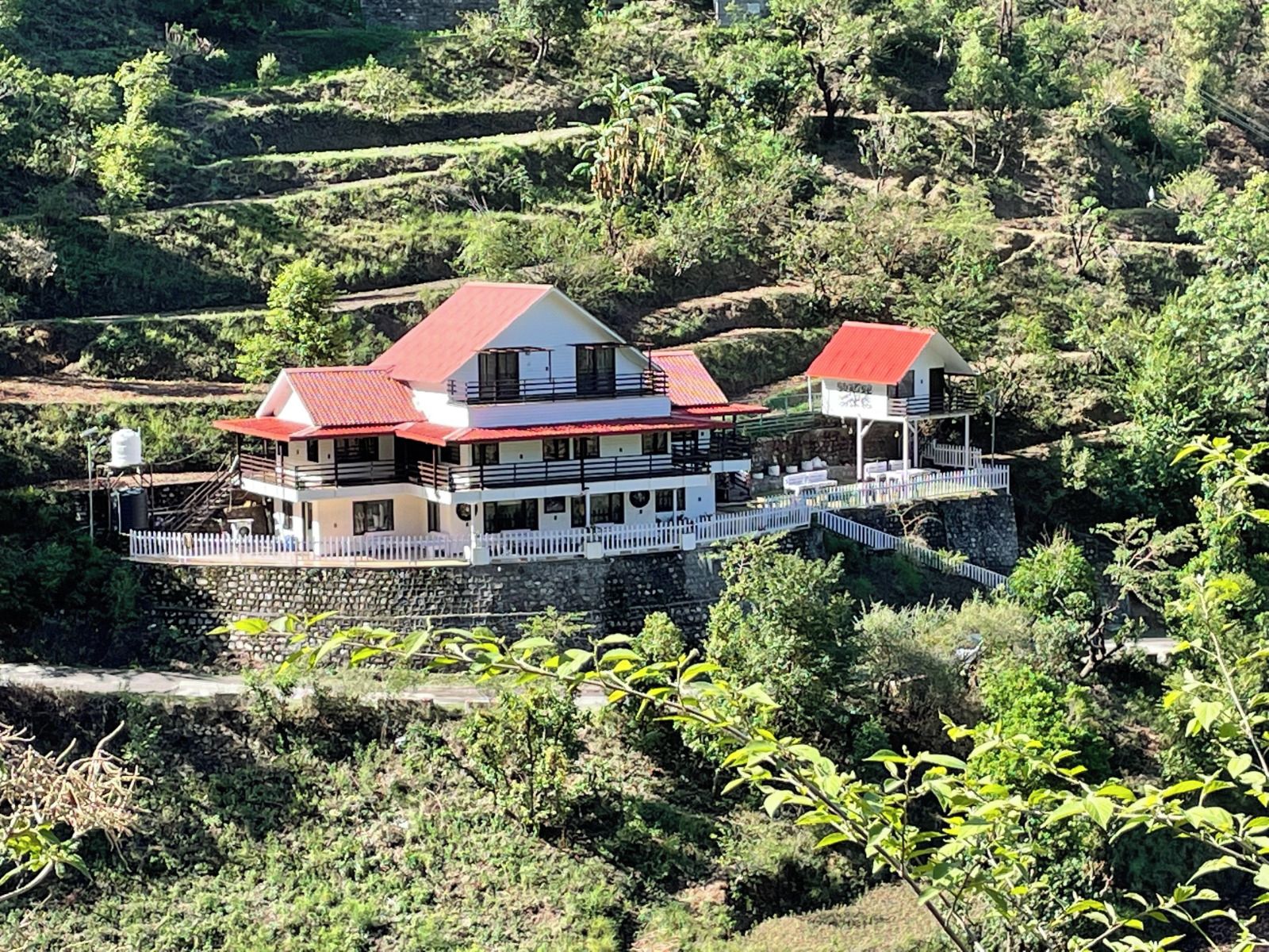 A two-story building with a red roof, situated on a terraced hillside with lush greenery.