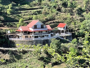 A two-story building with a red roof, situated on a terraced hillside with lush greenery. - Madhuram Luxury Villa (Dhanaulti)
