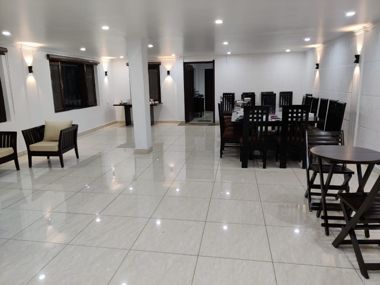 A spacious room with a tiled floor, a dining area with chairs and tables, and wall-mounted lighting.