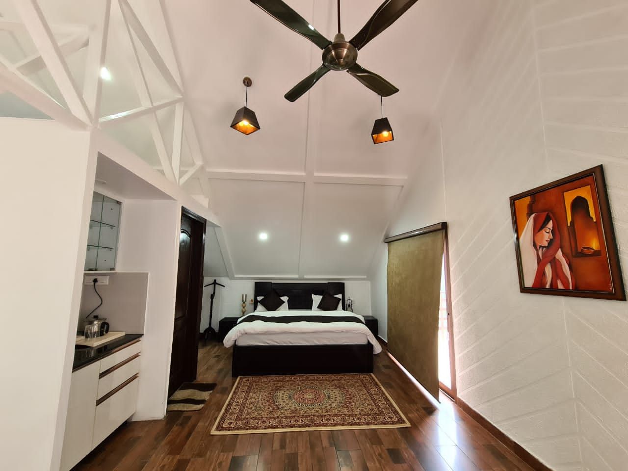 A bedroom with a bed, a ceiling fan, pendant lights, a painting, and a kitchen area with cabinets and a countertop.