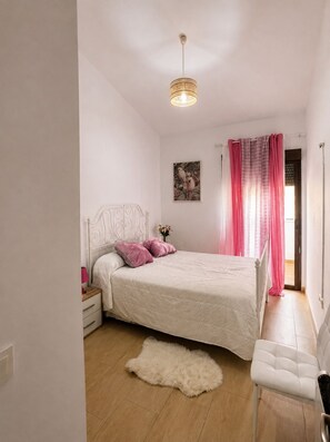 A bedroom with a bed, a nightstand, a chair, a fluffy rug, a hanging light fixture, a window with pink curtains, and a wall art piece. - Apartment in El Rompido with a pool | 3 bedrooms | 3 bathrooms (El Rompido)