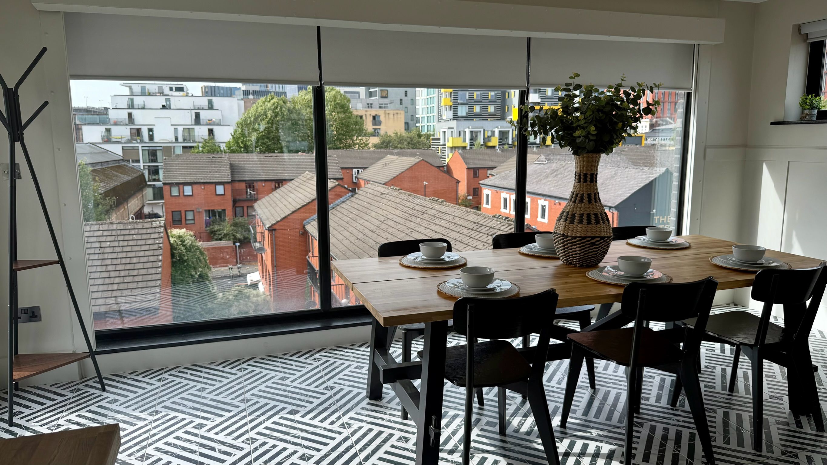 Un comedor con una mesa preparada para cuatro personas, un ventanal con vista a edificios y un suelo estampado.