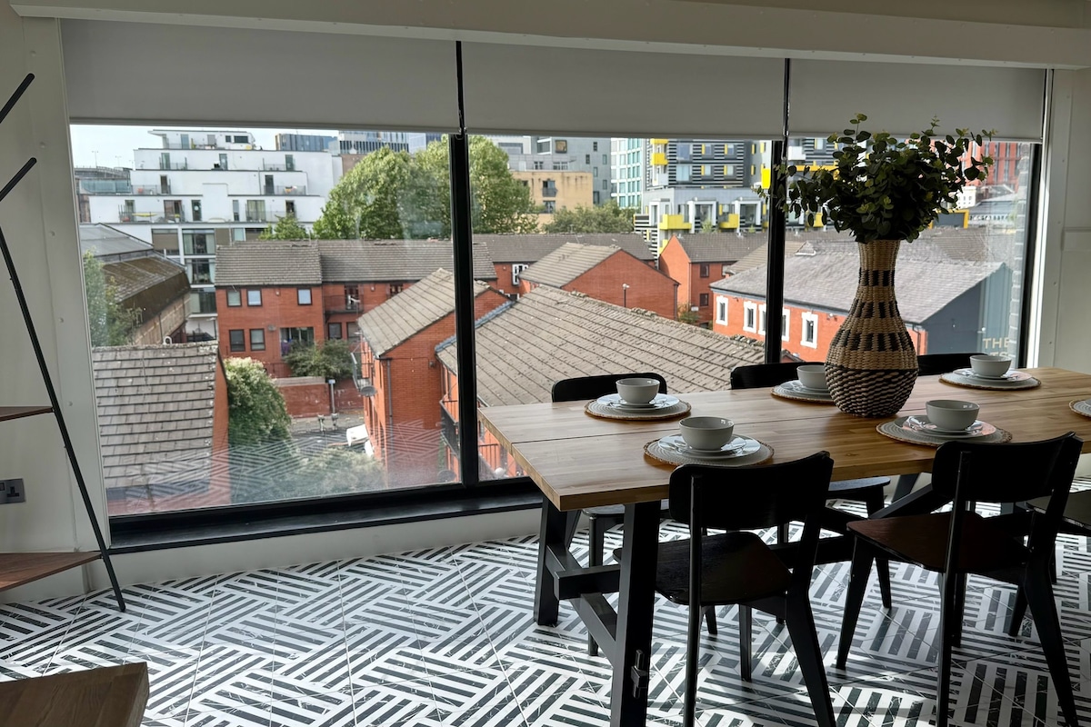 Un comedor con una mesa preparada para cuatro personas, un ventanal con vista a edificios y un suelo estampado.