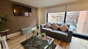 A modern living room with a grey sofa, a glass coffee table, and a large window offering a view of a brick building. - Celestial Apartments (Manchester)