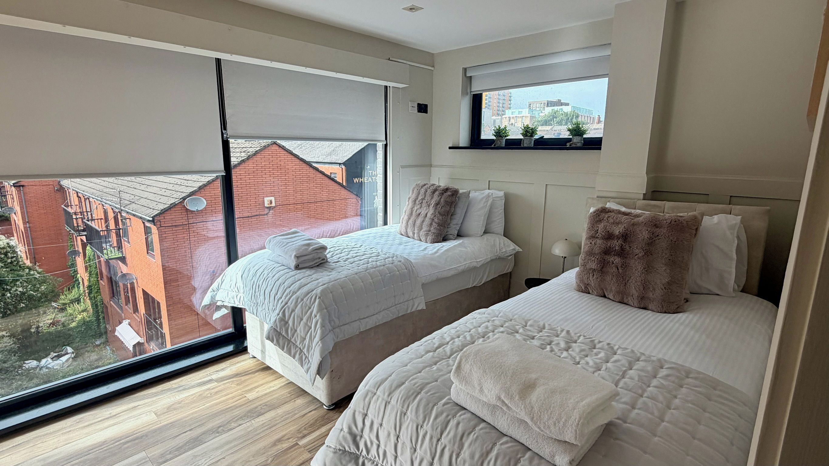 A bedroom with two beds, a window with a view of buildings, and a small shelf above the window.