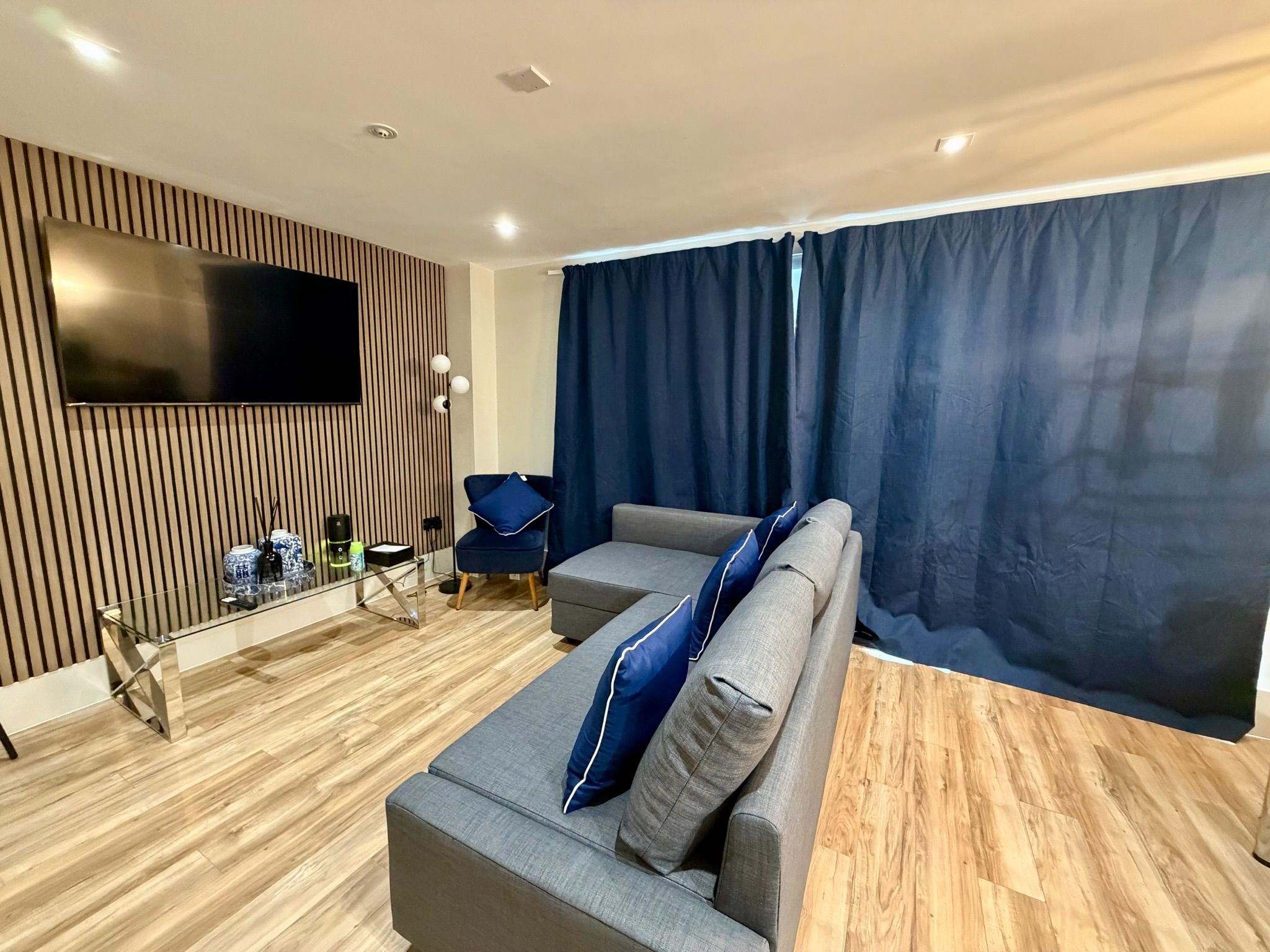 A modern living room with a flat-screen TV mounted on a wooden panel wall, a grey sofa with blue cushions, a glass coffee table, and a blue chair.