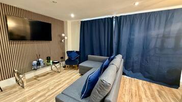 Una sala de estar moderna con un televisor de pantalla plana montado en una pared de paneles de madera, un sofá gris con cojines azules, una mesa de centro de vidrio y una silla azul.
