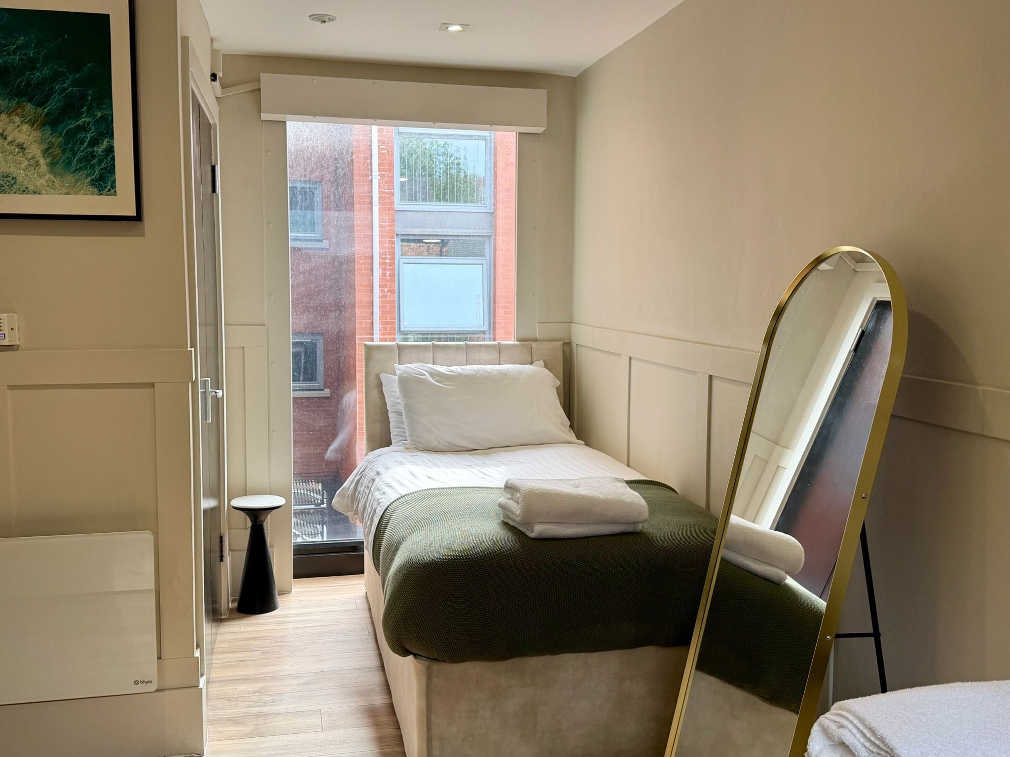 A hotel room with a bed, a small stool, a mirror, and a window with a view of a building.