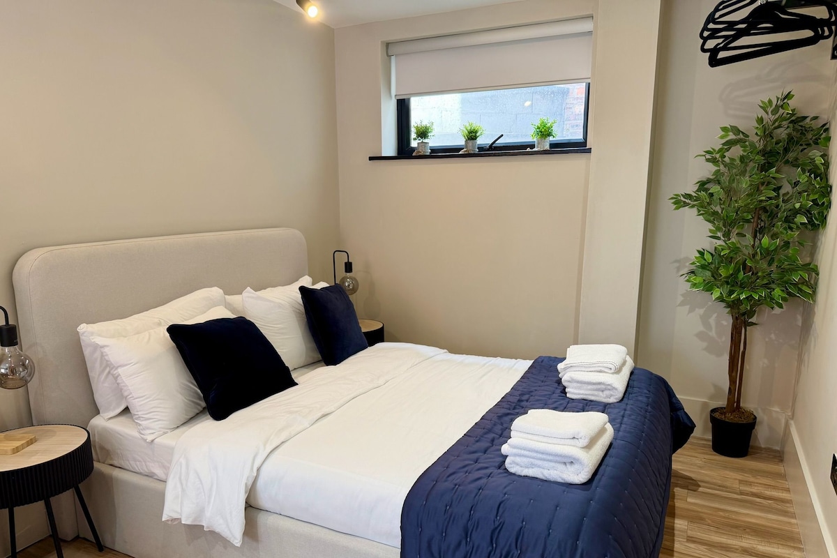 Una cama bien hecha con ropa de cama blanca y azul marino, una lámpara de noche y una ventana con plantas en macetas.
