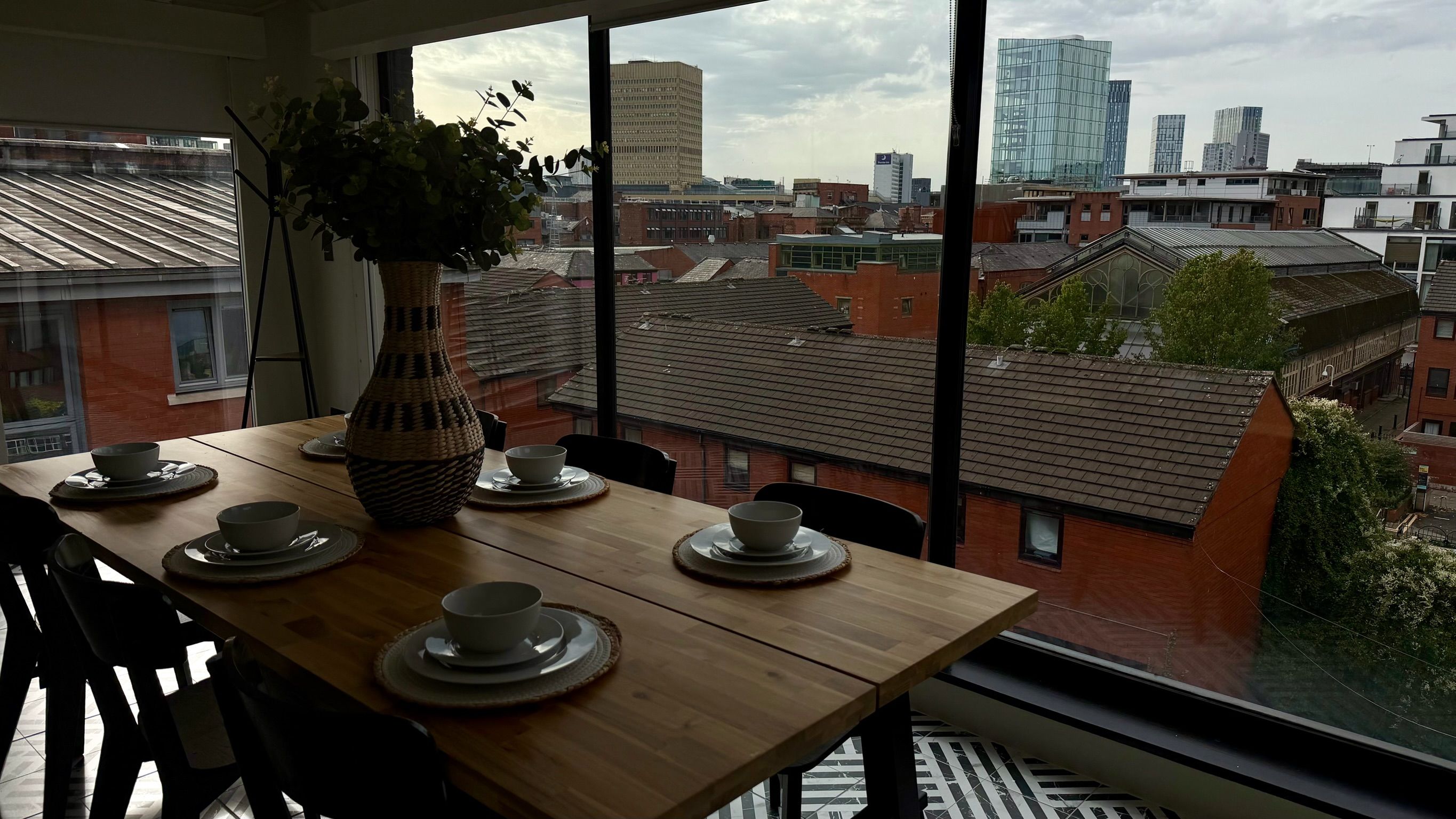 A dining table with plates and bowls, a vase, and a view of a cityscape through large windows.