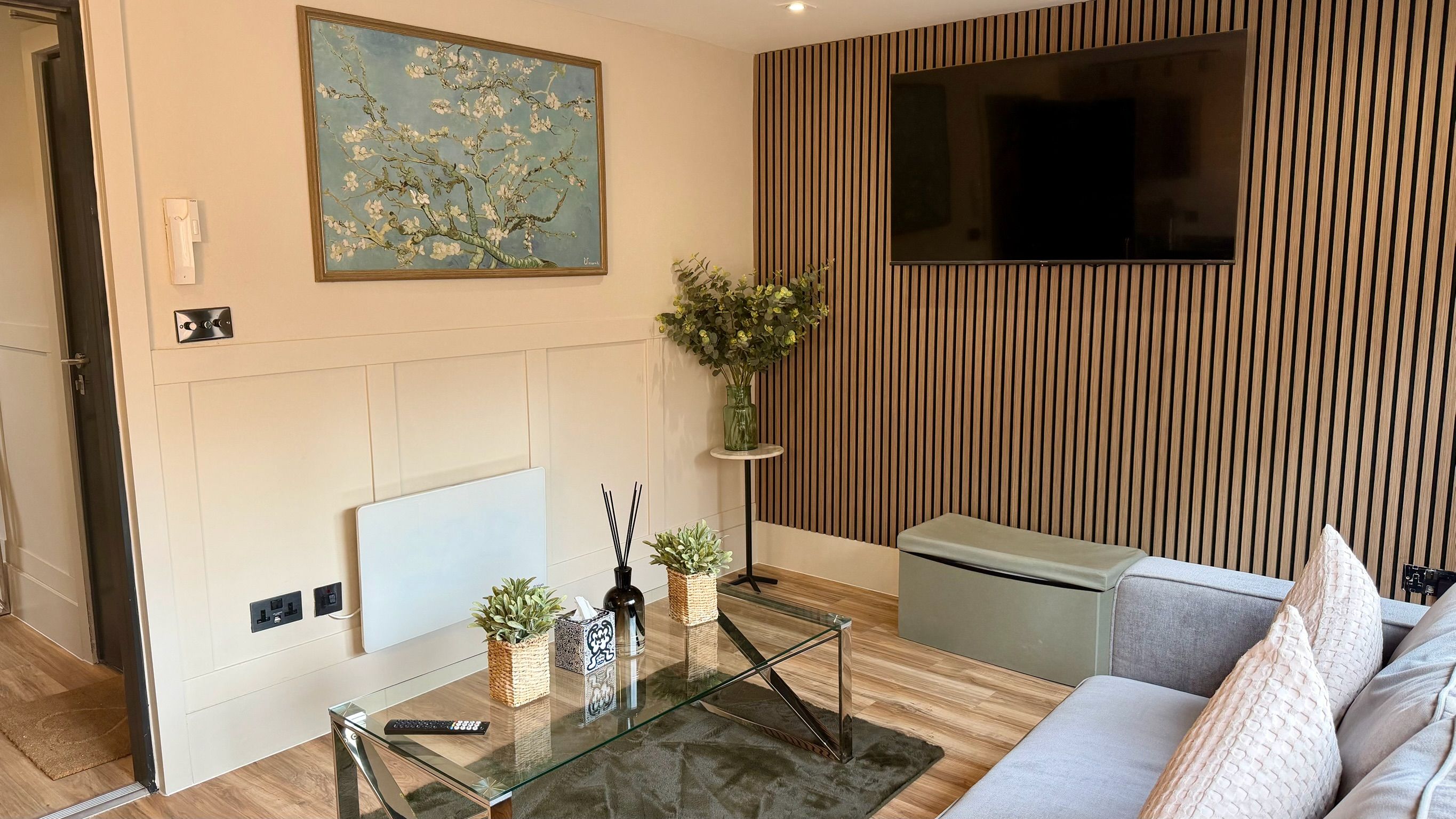 Una sala de estar moderna con chimenea, un televisor montado en una pared de madera, una mesa de centro de vidrio con controles remotos y un sofá con cojines.