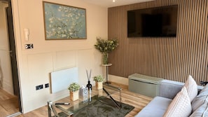 A modern living room with a fireplace, a television mounted on a wooden wall, a glass coffee table with remote controls, and a sofa with cushions. - Celestial Apartments (Manchester)