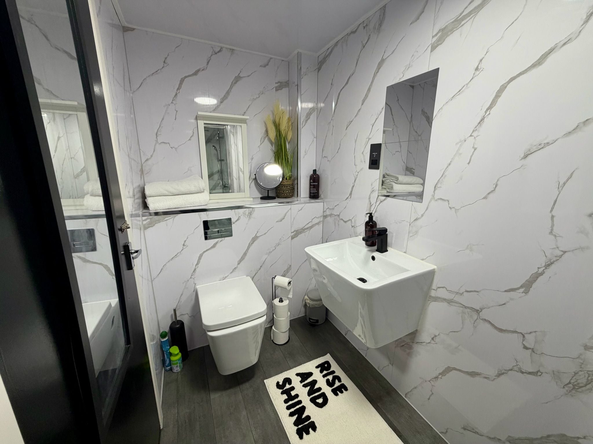 A bathroom with a white marble wall, a white sink, a toilet, and a mirror. There is a towel on the sink and a bottle of shampoo on the wall.
