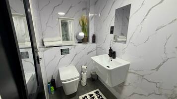 Un baño con pared de mármol blanco, un lavamanos blanco, un inodoro y un espejo. Hay una toalla sobre el lavamanos y una botella de champú en la pared.