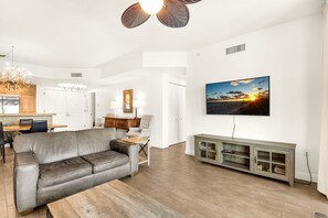 A modern living room with a grey sofa, wooden flooring, a dining area with a chandelier, and a TV mounted on the wall. - Portofino 2-1508 🌊🎾 Sound Views | Spa + Tennis + Fitness Onsite (Pensacola Beach)