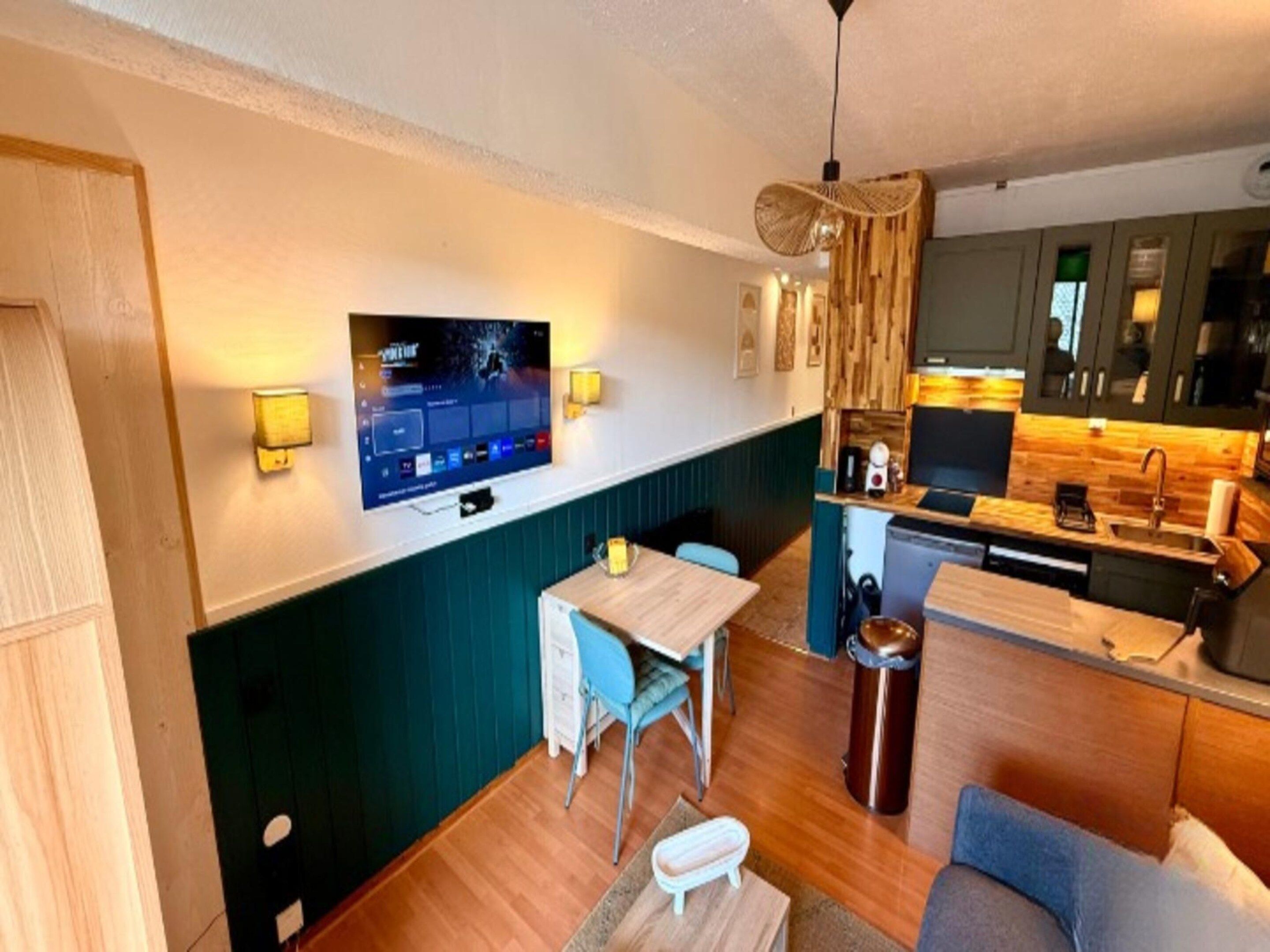 A compact living space with a kitchenette, a dining area with a table and chairs, a television mounted on the wall, and a trash bin.