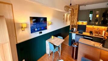 A compact living space with a kitchenette, a dining area with a table and chairs, a television mounted on the wall, and a trash bin.