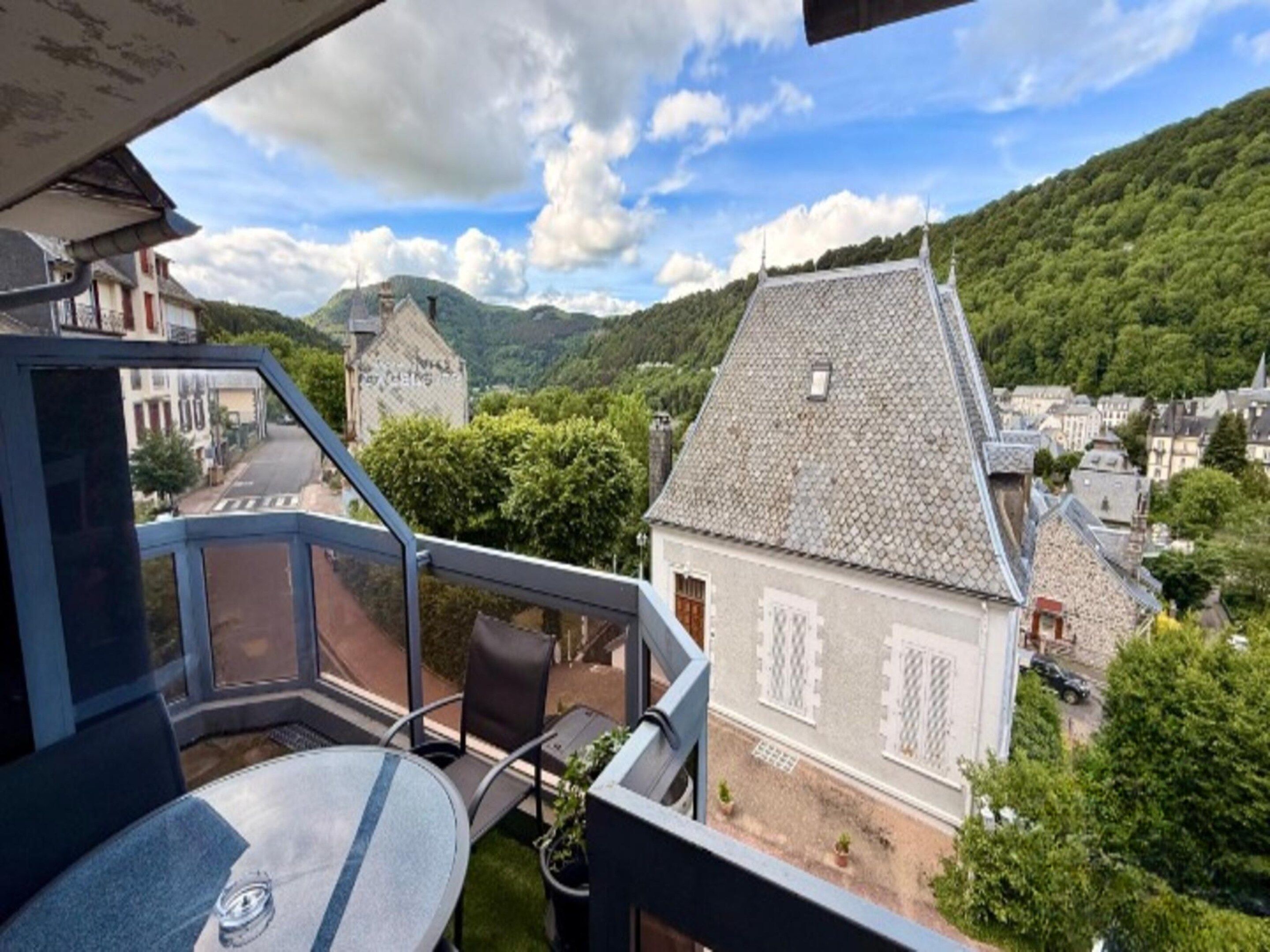 A balcony with a table and chairs, overlooking a village with traditional buildings and a mountainous backdrop.