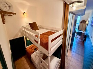 Quarto com beliche, uma pequena escrivaninha e uma cadeira, luminária na parede e vista para um corredor.