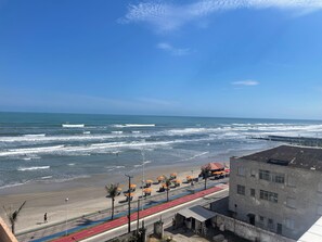 A coastal road with a beach on one side, a pedestrian path, and a building with a red roof. - 2 bedroom apartment, 1 suite, perfect location, new building, beachfront, downtown. (Mongaguá)