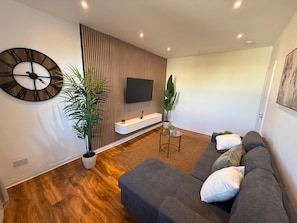 A modern living room with a sectional sofa, a large wall clock, a flat-screen TV, and a wooden accent wall. - Luxury 1-Bed, Free Parking, 15 Mins Central London, Sleeps 4 (London)