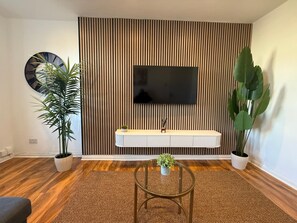 A modern living room with a flat-screen TV mounted on a wooden slat wall, a white console table, a round glass coffee table, and two potted plants. - Luxury 1-Bed, Free Parking, 15 Mins Central London, Sleeps 4 (London)