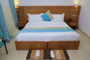 Een tweepersoonsbed met houten hoofdbord, twee nachtkastjes, een nachtstandaard en een paar slippers op een kleed.