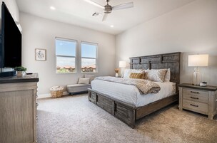 Quarto com cama grande, mesa de cabeceira, cômoda, ventoinha no teto e janela com vista para casas.