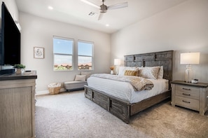 Quarto com cama grande, mesa de cabeceira, cômoda, ventoinha no teto e janela com vista para casas.