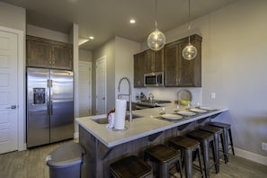 A modern kitchen with a stainless steel refrigerator, a built-in microwave, a large island with bar stools, and pendant lighting. - Snow Canyon Escape Private Pool #65 5 Bedroom Townhouse (Santa Clara)