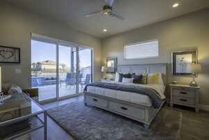 Quarto com cama grande, mesa de vidro, cadeira, mesa de cabeceira e ventilador de teto.