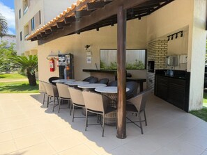 An outdoor patio with a brick fireplace, wicker furniture, and a tiled floor. - Furnished 2-bedroom apartment in Ponta Negra, condominium with pool, close to the beach. (Natal)