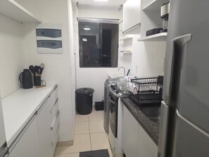 A compact kitchen with a refrigerator, stove, sink, and a window above the sink. - Furnished 2-bedroom apartment in Ponta Negra, condominium with pool, close to the beach. (Natal)