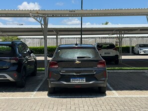 A parking area with a covered structure, a car with a visible license plate, and a building with a metal roof in the background. - Furnished 2-bedroom apartment in Ponta Negra, condominium with pool, close to the beach. (Natal)