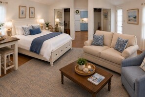 A well-lit bedroom with a bed, bedside tables, and a nightstand. There is a living area with a sofa, armchair, and coffee table. The room has a neutral color palette and includes a kitchen area visible through an open door. - Petite downtown studio cottage (Havre de Grace)