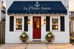 A white building with a red door and black shutters, labeled 'La Petite Ancre Vacation Rental.' - Petite downtown studio cottage (Havre de Grace)