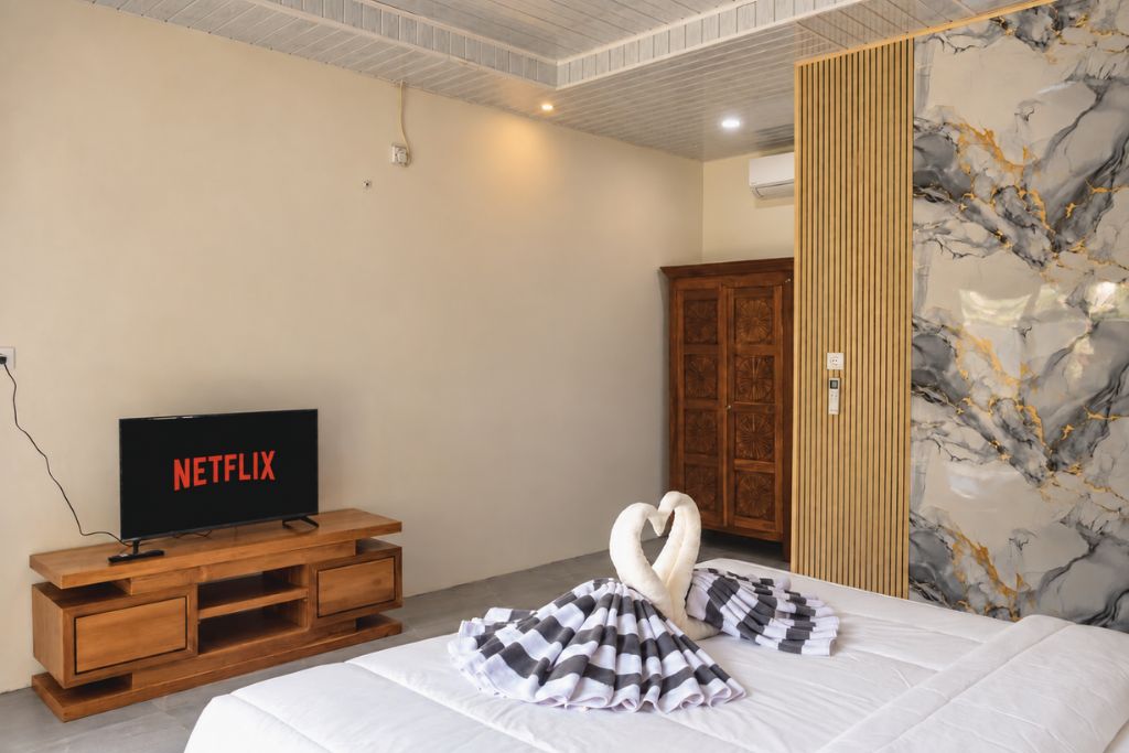 Ein Hotelzimmer mit einem Bett, einer hölzernen Kommode, einem Fernseher, der 'NETFLIX' anzeigt, einer herzförmigen Dekoration auf dem Bett und einer Wand mit Marmormuster.