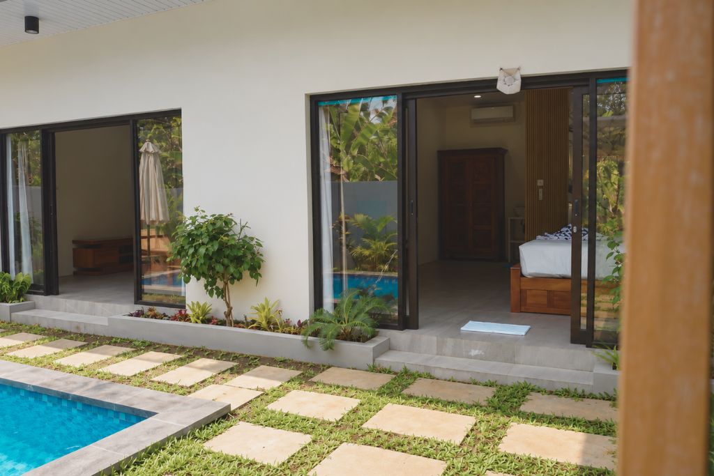 Zona de piscina exterior moderna amb entrada de porta de vidre, un petit jardí amb camins pedrestres i una cadira de fusta.