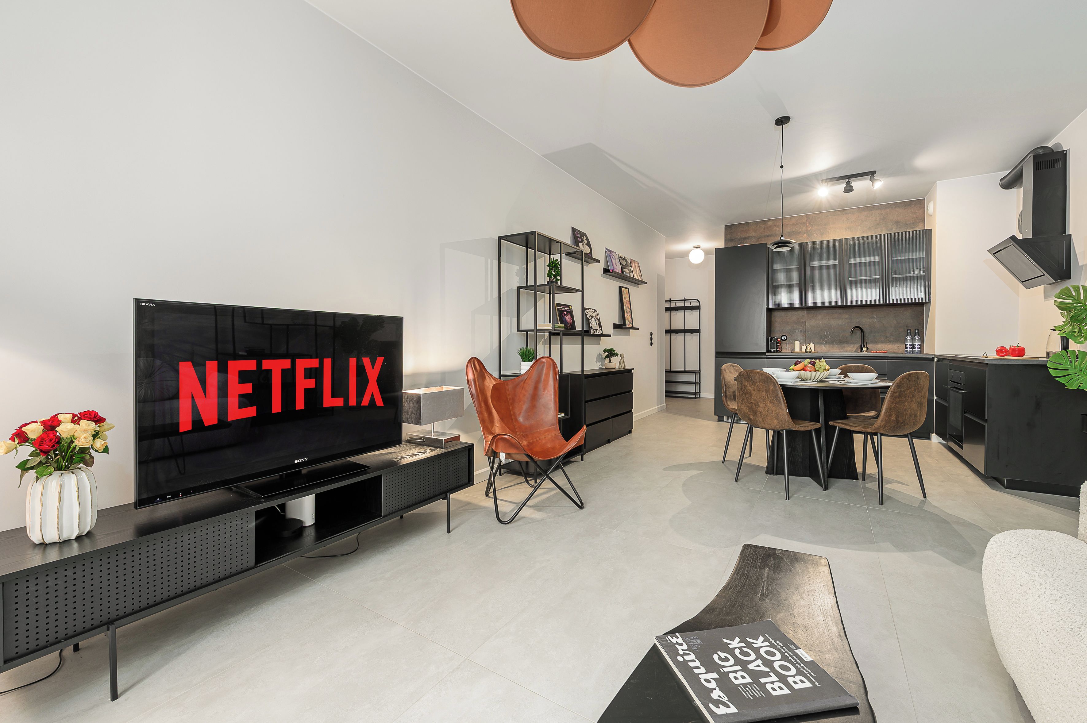 A modern living room with a flat-screen TV displaying 'NETFLIX,' a dining area with a round table, and a kitchen with a built-in oven and microwave.
