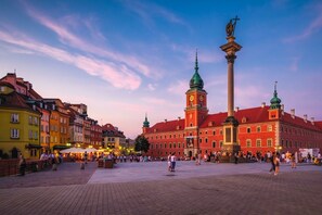 A historic square with a prominent red building featuring a clock tower and two green spires, surrounded by colorful buildings and a paved area with people. - Lumina spacious Apart Parking I Terrace (Warszawa)