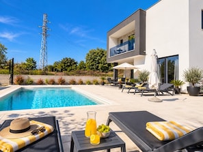 Zona de piscina exterior moderna amb gandules, una taula amb una beguda i un edifici amb un balcó.