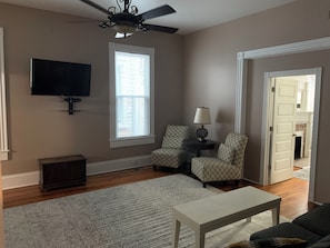 A living room with a ceiling fan, a flat-screen TV, patterned chairs, a lamp, and a white coffee table. - That big blue house downtown! (Wilmington)