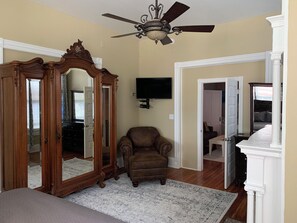 A room with a ceiling fan, a large mirror, a brown armchair, a television, and a bed with a white headboard. - That big blue house downtown! (Wilmington)