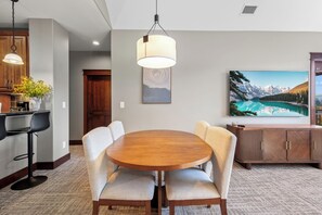 A dining area with a round wooden table, four upholstered chairs, a hanging light fixture, a wall-mounted painting, and a kitchen area with wooden cabinets and a potted plant. - New Listing! Riverstone Condo | Private Balcony | Walk to Shops and Dining (Coeur d'Alene)