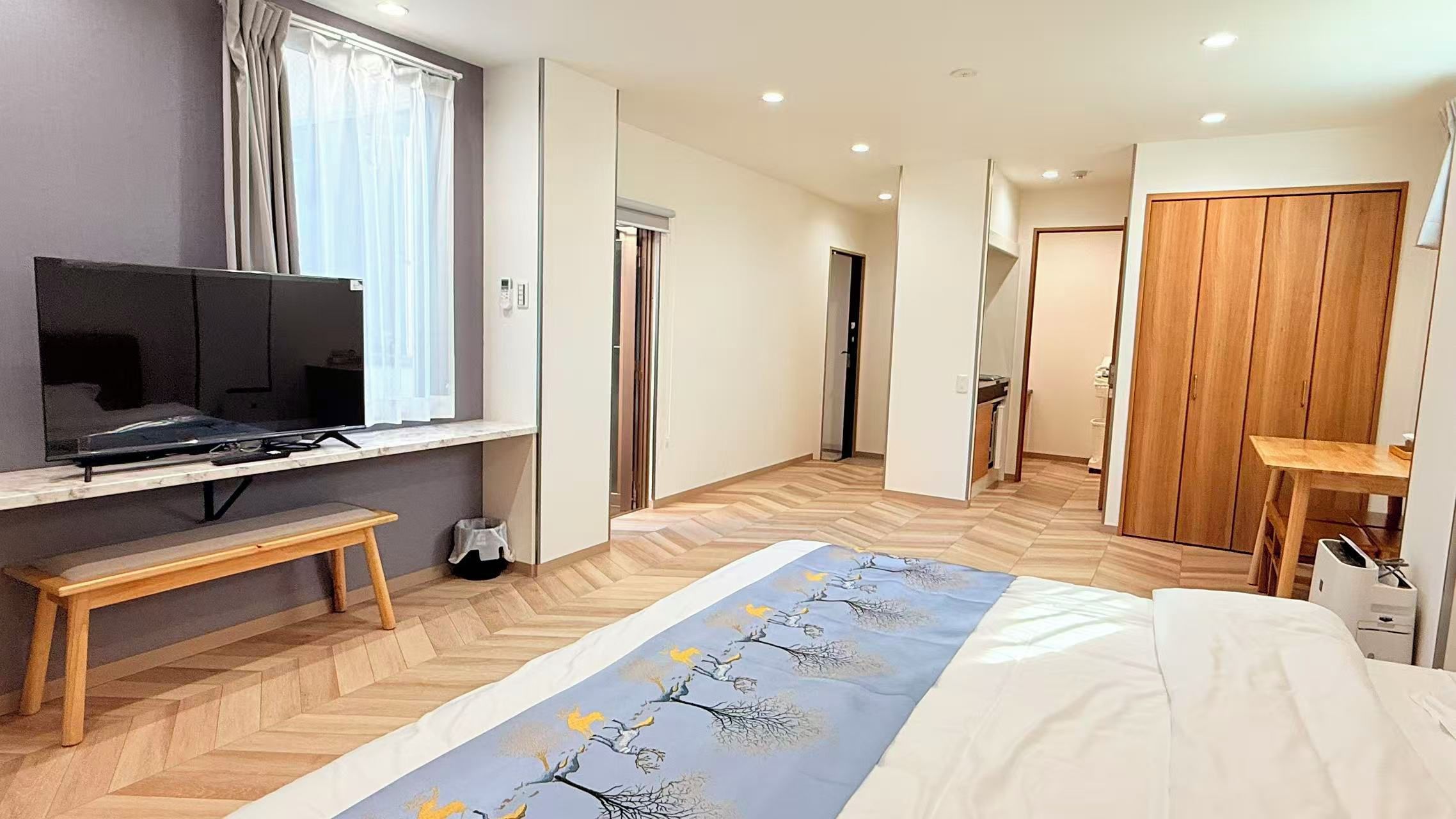 A hotel room with a bed, a TV on a stand, a wooden bench, a desk, and a wardrobe.