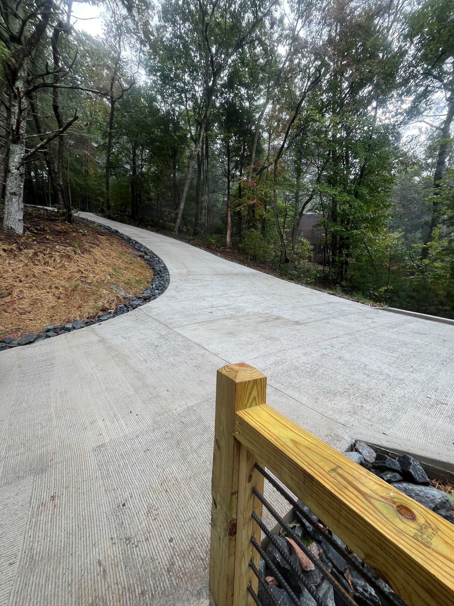 A paved pathway curving through a wooded area with a wooden fence on one side.