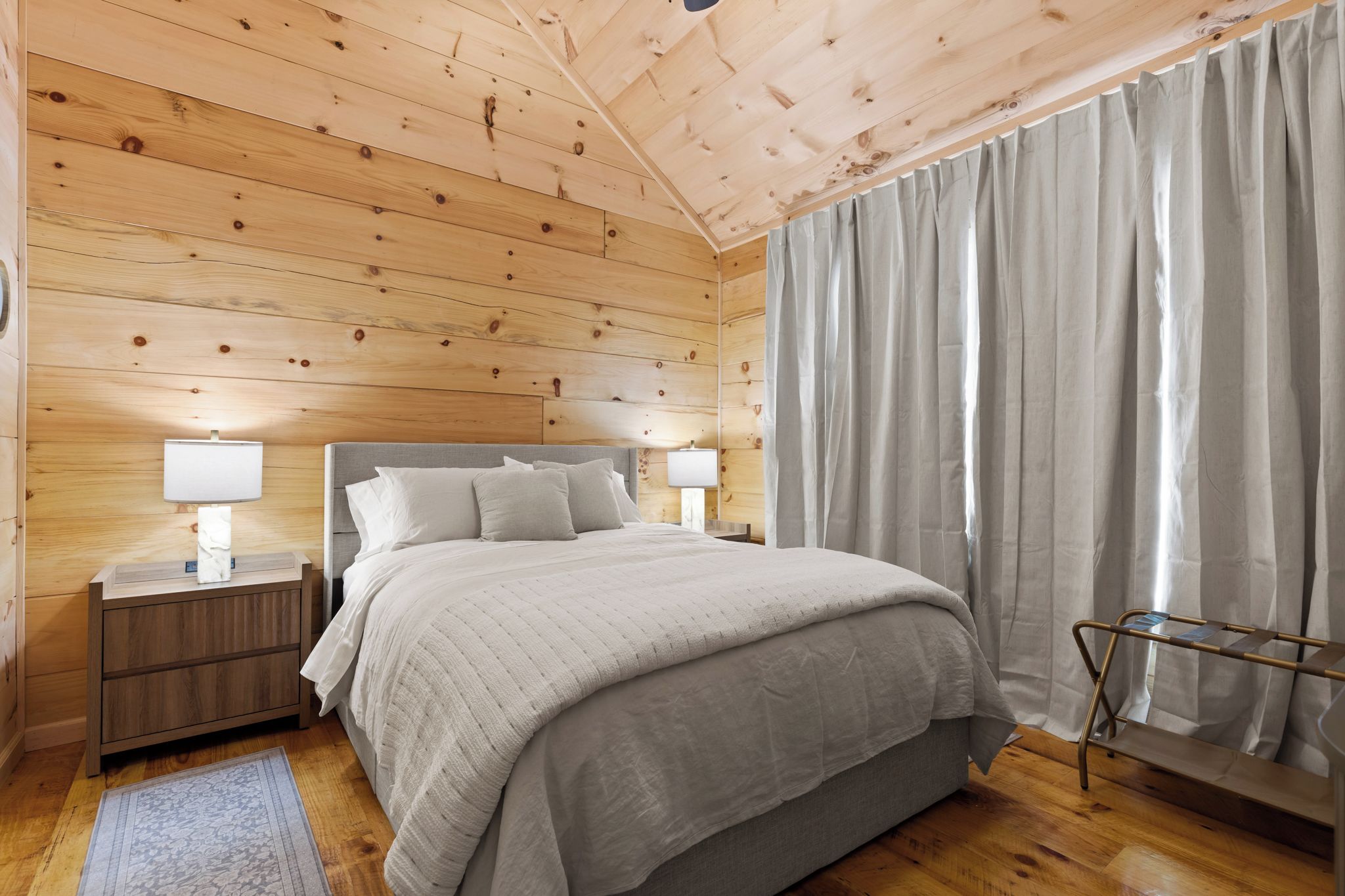 Un dormitorio con paneles de madera, una cama, mesitas de noche y un ventanal con cortinas.
