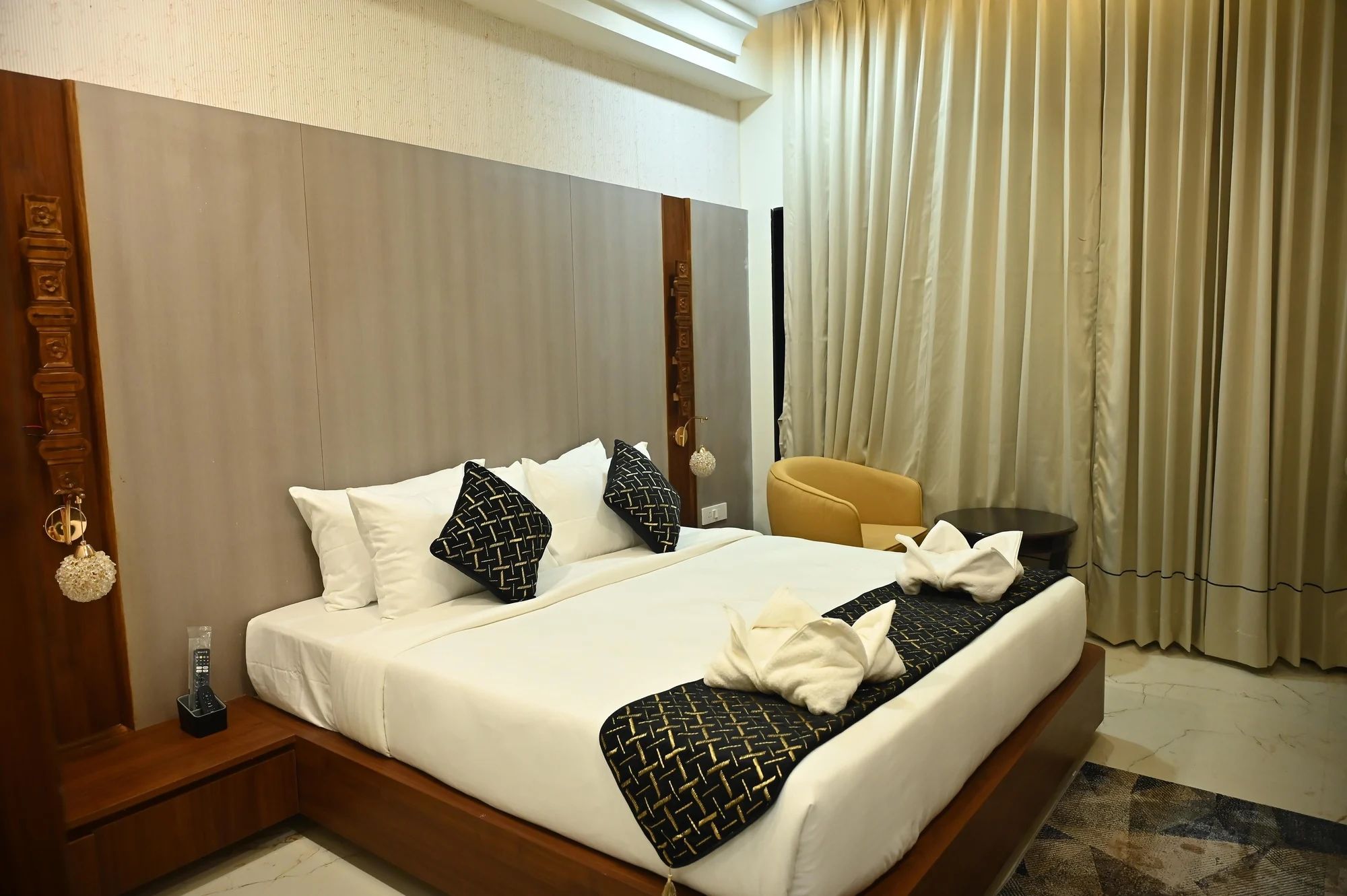 A hotel room with a large bed, a wooden headboard, a bedside table with a remote control, a yellow armchair, and a window with curtains.